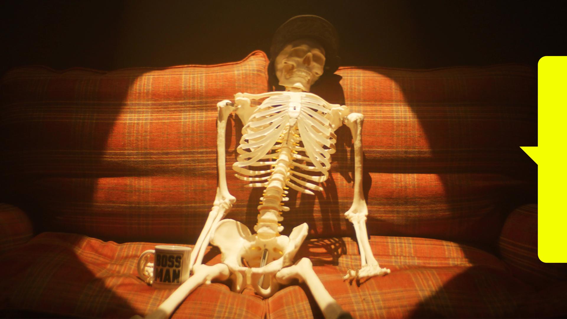 Ein menschliches Skelett sitzt in einem Lichtkegel auf einem karierten Sofa. Es trägt eine Mütze und hat eine Tasse neben sich stehen.