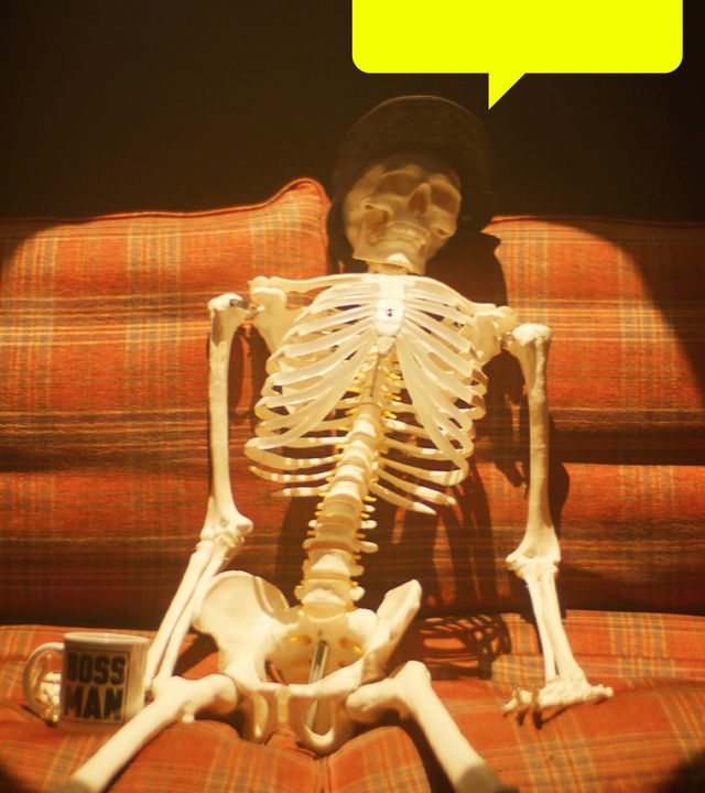 Ein menschliches Skelett sitzt in einem Lichtkegel auf einem karierten Sofa. Es trägt eine Mütze und hat eine Tasse neben sich stehen.