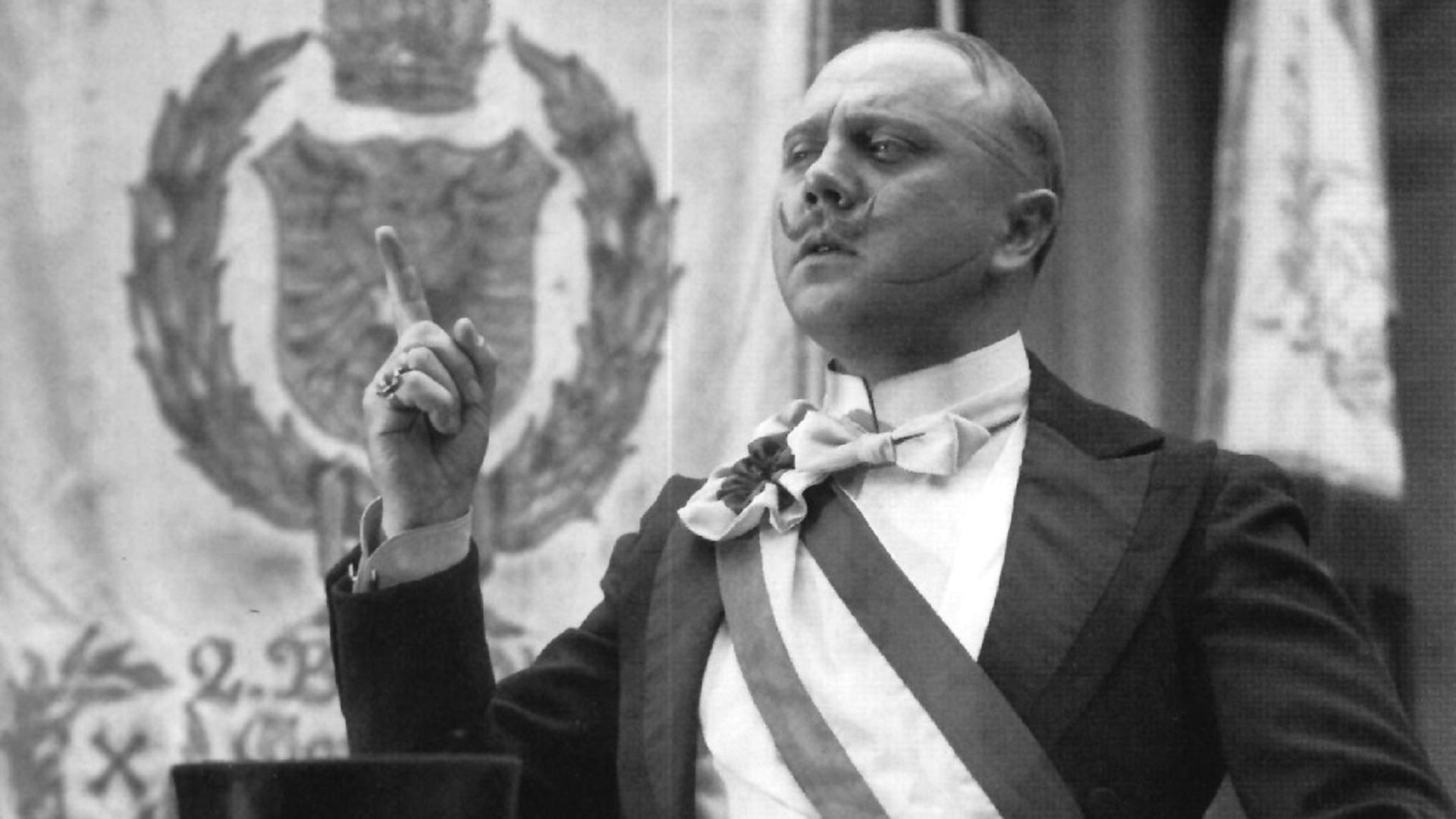 Diederich Heßling (Werner Peters) steht mit hochgezwirbeltem Schnurrbart hinter einem Rednerpult und reckt mit selbstgewissem Blick den Zeigerfinger. Er trägt ein festliches schwarzes Jackett und ein weißes Hemd mit Stehkragen, darüber eine dreifarbige Schärpe. Vor ihm auf dem Pult sind ein schwarzer Zylinder und weiße Handschuhe abgelegt. Im Hintergrund ist eine Fahne mit Kaiserwappen aufgehängt.