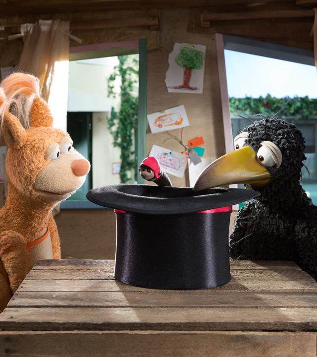  Eichhörnchen Ella und Rudi sind im Baumhaus und schauen neugierig auf den Zylinder vor ihnen auf dem Tisch. Oben auf der Krempe liegt ein kleines Plüschkaninchen, auf dem Tisch vor Rudi ein Zauberstab.