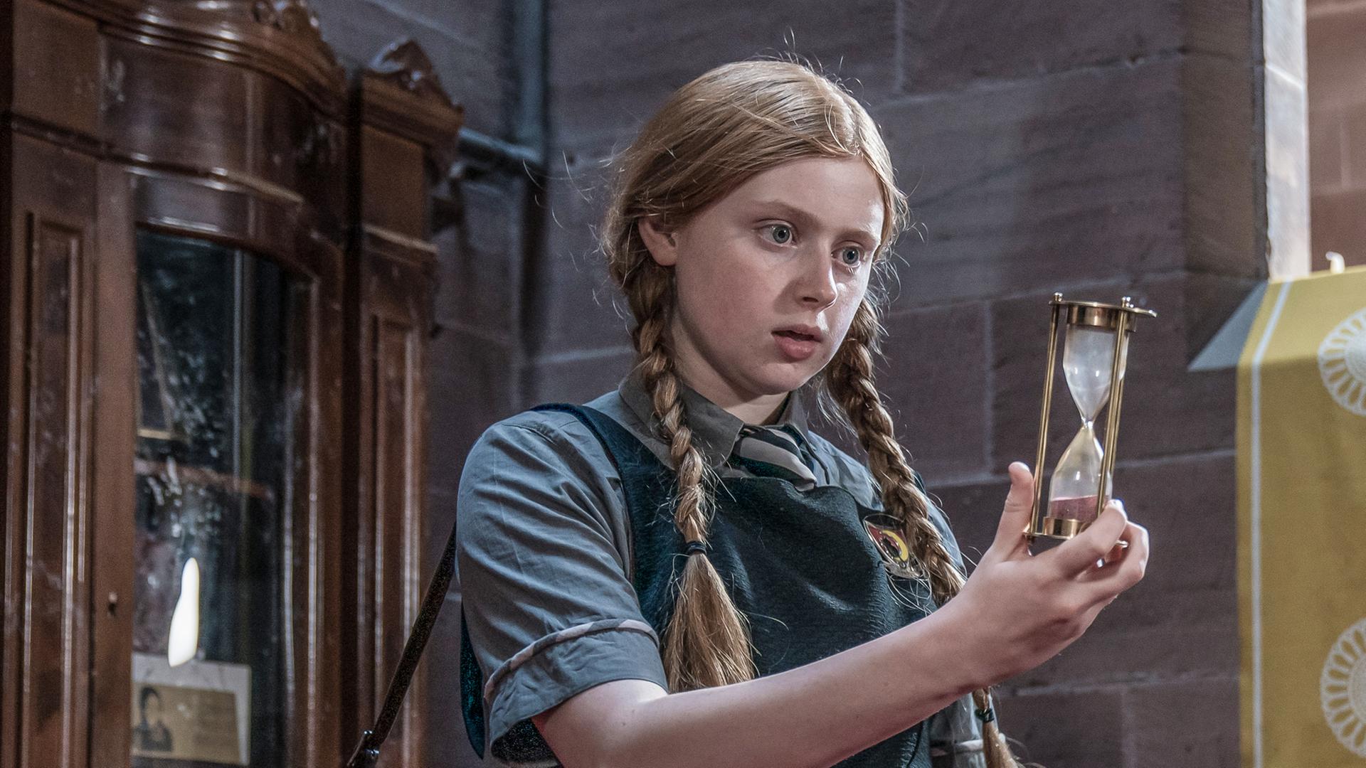 Der Zeitzauber: In einer Halle innerhalb der Grausteinakademie steht Mildred in Schuluniform und Schultasche. Sie hält ein Stundenglas vor sich in die Hand und schaut darauf. Der Sand im Stundenglas befindet sich unten am Boden.