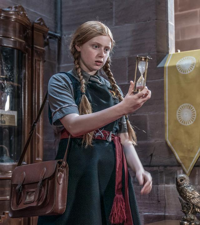 Der Zeitzauber: In einer Halle innerhalb der Grausteinakademie steht Mildred in Schuluniform und Schultasche. Sie hält ein Stundenglas vor sich in die Hand und schaut darauf. Der Sand im Stundenglas befindet sich unten am Boden.