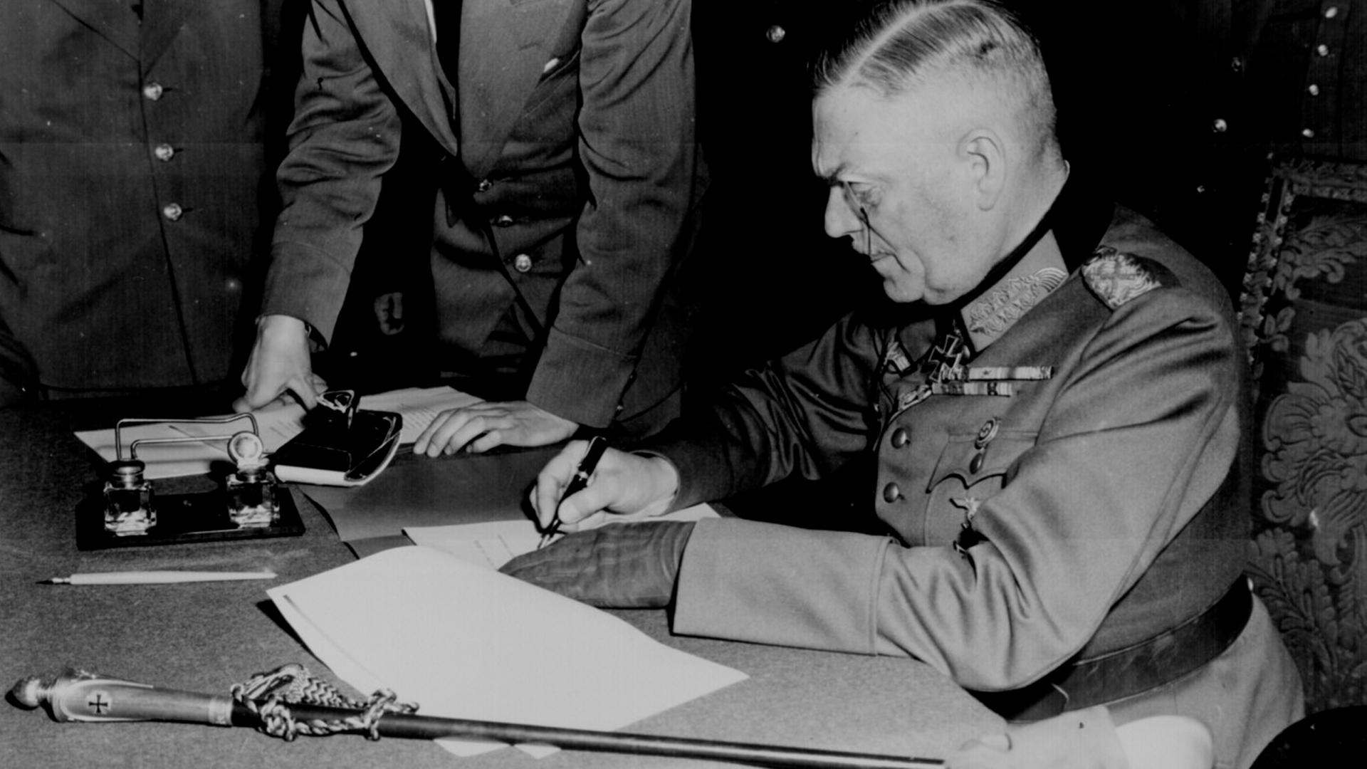 "Der Zweite Weltkrieg: Apokalypse": Schwarz-Weiß-Aufnahme: Generalfeldmarschall Wilhelm Keitel sitzt an einem Tisch und unterschreibt die Kapitulation der Wehrmacht.