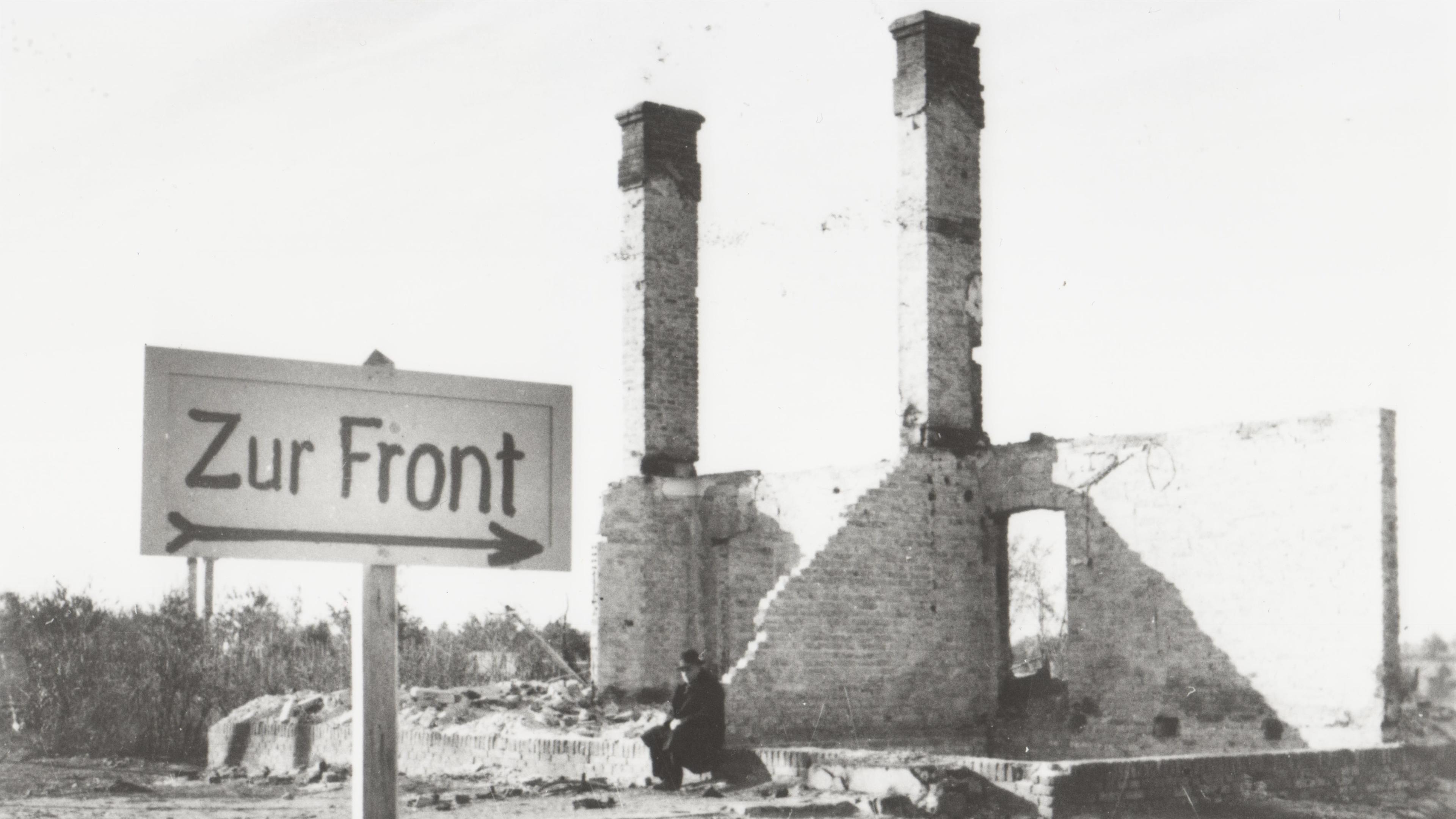 "Der Zweite Weltkrieg: Der Überfall": Ein Mann sitzt in den Ruinen eines durch den Krieg zerstörten Hauses in einer polnischen Ortschaft. Im Vordergrund ist ein Wegweiser mit der Aufschrift "Zur Front" errichtet worden.