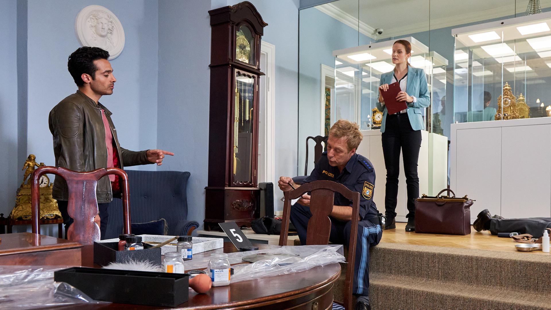 "Die Rosenheim-Cops - Des Kaisers letzte Stunde": Kommissar Kaya (Baran Hêvî) steht in einem Raum hinter einem antiken dunklen Holztisch, um den Stühle herumstehen, und zeigt auf Polizeihauptmeister Mohr (Max Müller), der auf einem dreistufigen Treppenabsatz sitzt. Neben ihm steht eine antike Pendeluhr. Oberhalb des Treppenabsatzes steht Pathologin Mai (Sina Wilke), die eine Kladde in den Händen hält. Neben ihr steht eine große braune Tasche, hinter ihr sind Glasvitrinen mit weißen Sockeln zu sehen, in denen goldene Tischuhren stehen.