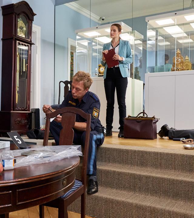 "Die Rosenheim-Cops - Des Kaisers letzte Stunde": Kommissar Kaya (Baran Hêvî) steht in einem Raum hinter einem antiken dunklen Holztisch, um den Stühle herumstehen, und zeigt auf Polizeihauptmeister Mohr (Max Müller), der auf einem dreistufigen Treppenabsatz sitzt. Neben ihm steht eine antike Pendeluhr. Oberhalb des Treppenabsatzes steht Pathologin Mai (Sina Wilke), die eine Kladde in den Händen hält. Neben ihr steht eine große braune Tasche, hinter ihr sind Glasvitrinen mit weißen Sockeln zu sehen, in denen goldene Tischuhren stehen.