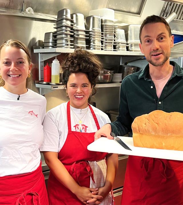 Drei Personen stehen in einer professionellen Küche und halten einen Kuchen