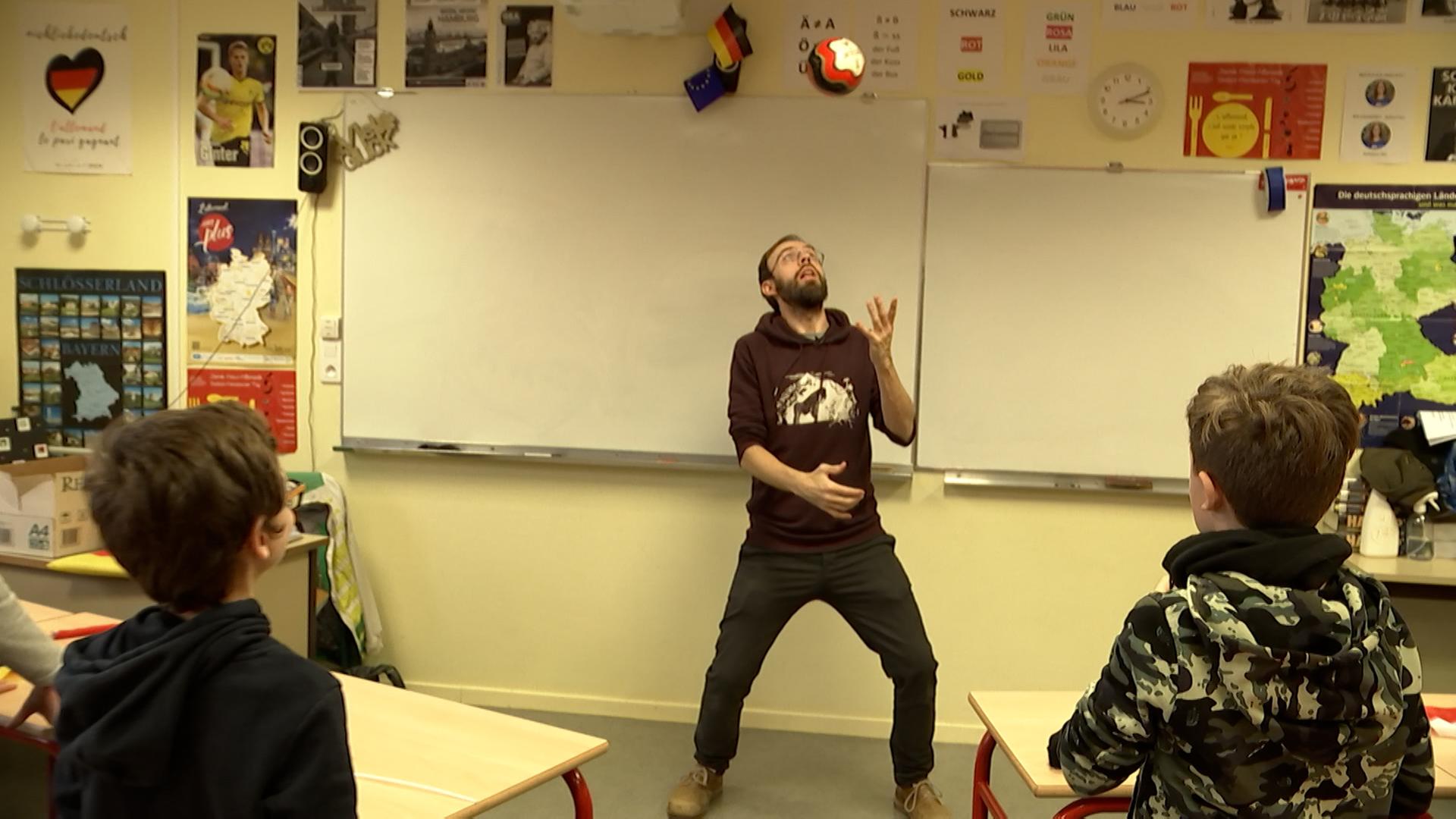 In einem Klassenzimmer macht ein Mann mit Bart einen Kopfball vor der Tafel. Im Hintergrund hägt eine kleine Deutschlandflagge. 