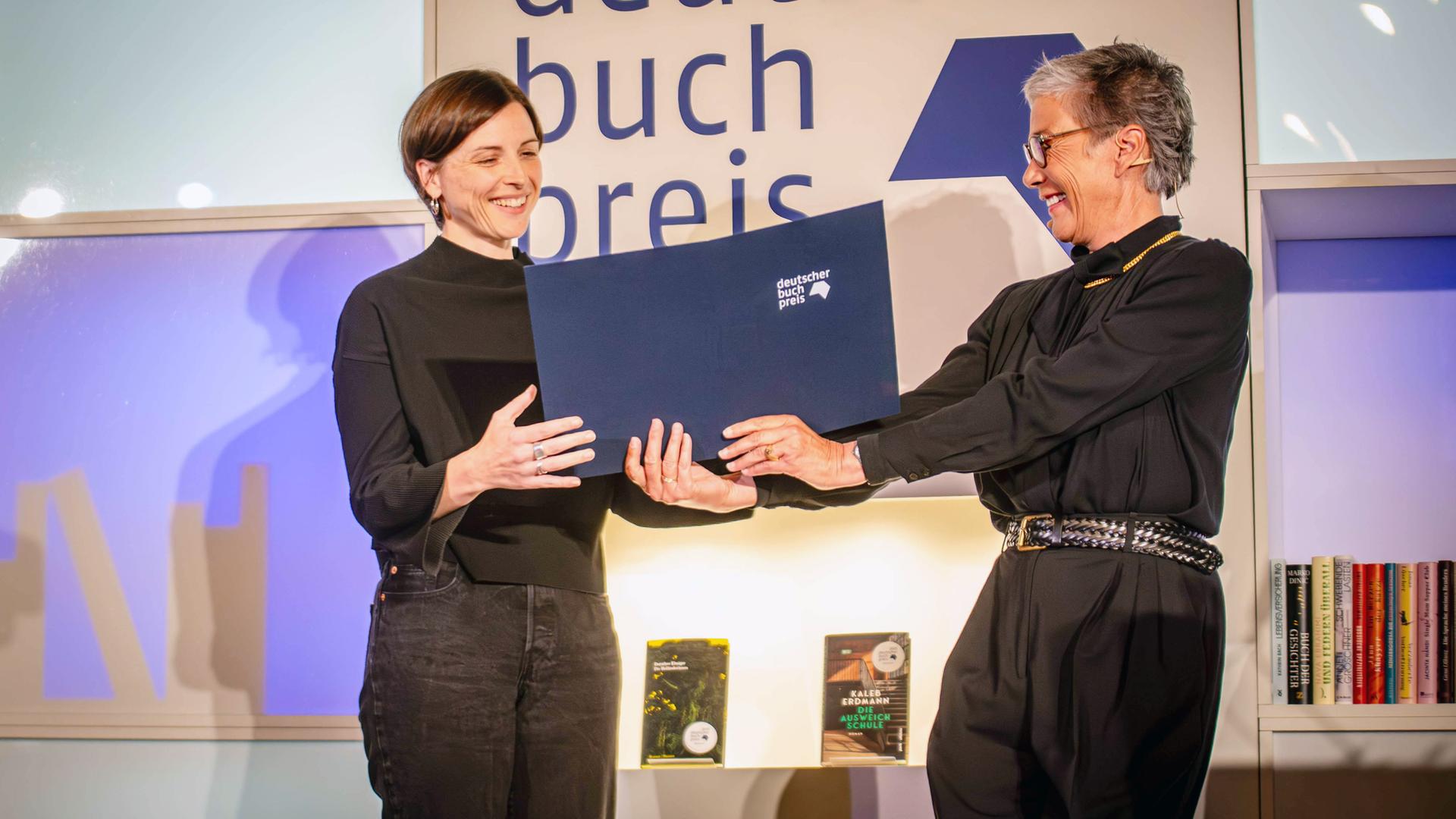 Eine schwarz gekleidete Frau mit graumeliertem Haar und Brille überreicht einer Frau mit kurzen braunen Haaren, die glücklich lächelt, eine blaue Kladde. Hinter ihnen steht in Großbuchstaben Deutscher Buchpreis
