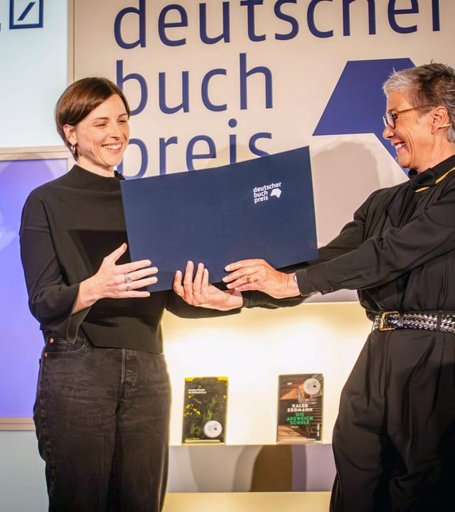Eine schwarz gekleidete Frau mit graumeliertem Haar und Brille überreicht einer Frau mit kurzen braunen Haaren, die glücklich lächelt, eine blaue Kladde. Hinter ihnen steht in Großbuchstaben Deutscher Buchpreis