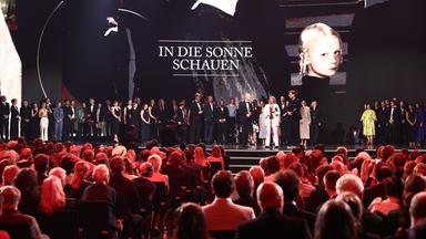 Das Team von "In die Sonne schauen" versammeln sich bei der Verleihung des Deutschen Filmpreises auf der Bühne. Hinter ihnen werden Filmszenen und der Titel eingeblendet, vor ihnen sitzt ein dicht besetztes Publikum im Saal.