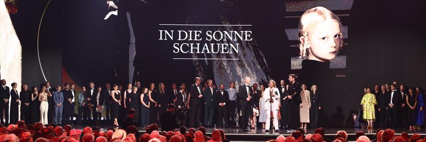 Das Team von "In die Sonne schauen" versammeln sich bei der Verleihung des Deutschen Filmpreises auf der Bühne. Hinter ihnen werden Filmszenen und der Titel eingeblendet, vor ihnen sitzt ein dicht besetztes Publikum im Saal.