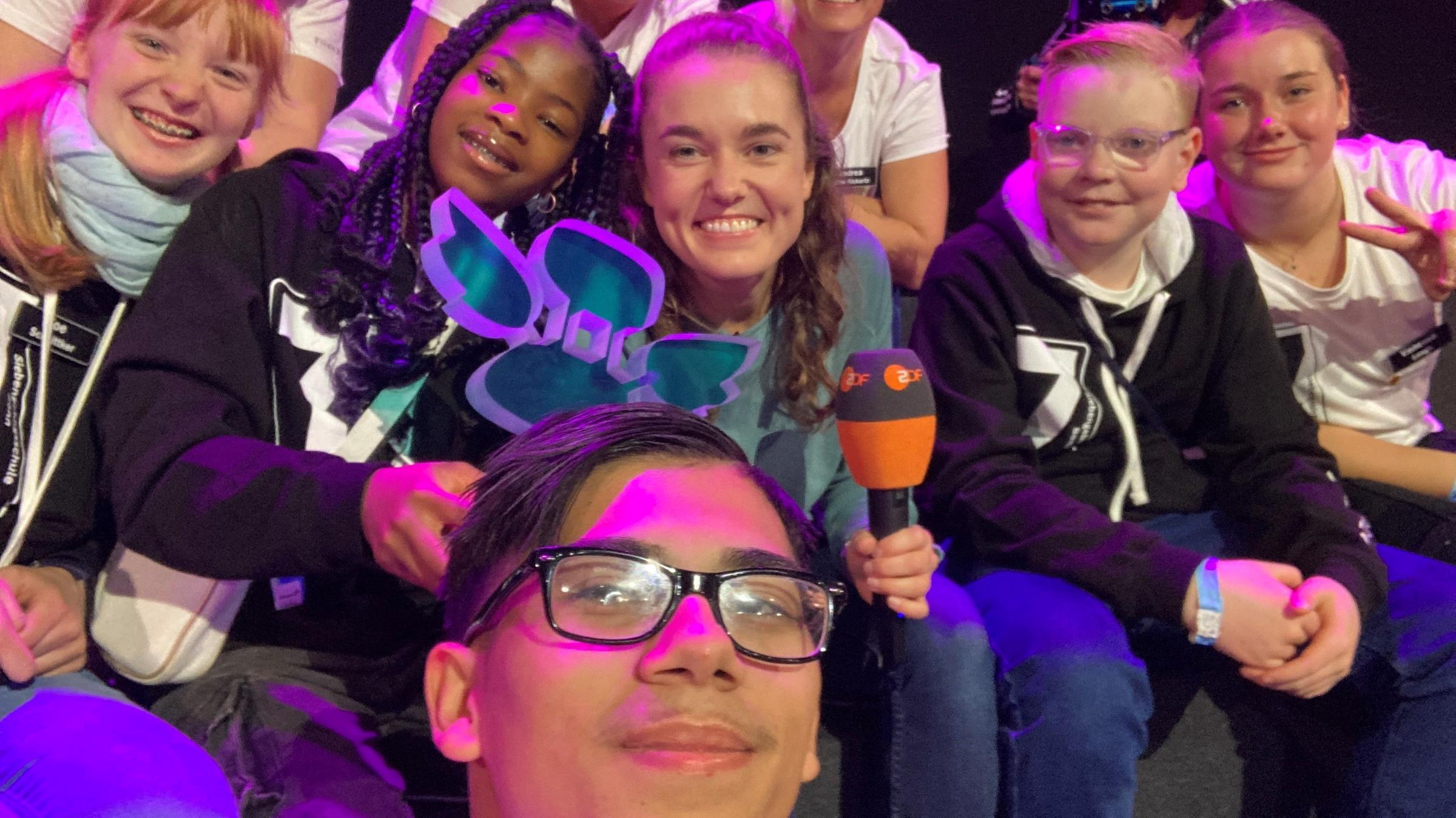 logo!-Moderatorin Lotte sitzt in der Mitte von Schülerinnen und Lehrerinnen