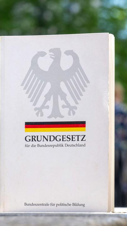Das deutsche Grundgesetz steht in einer Einkaufsstraße, im Hintergrund sind verschwommen Menschen zu sehen.