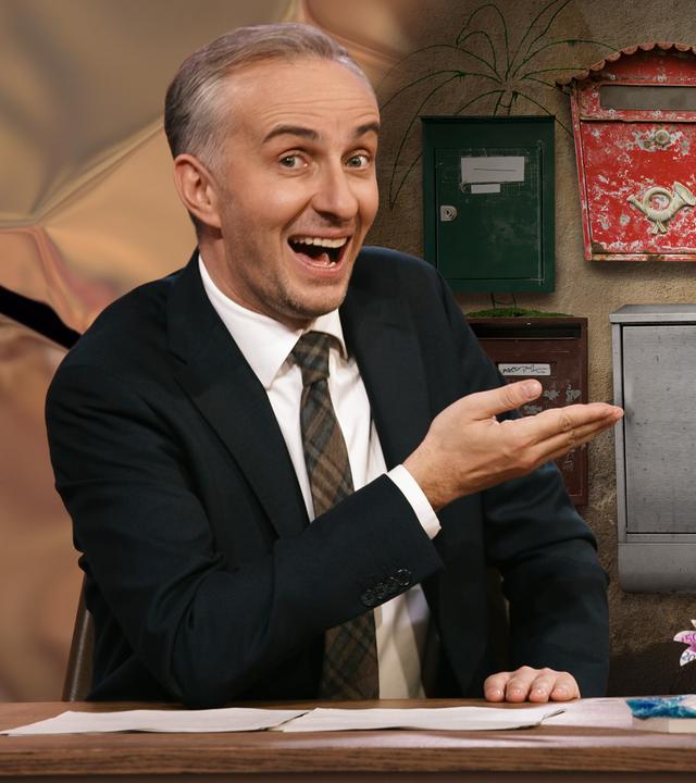 Jan Böhmermann sitzt am Schreibtisch und zeigt auf eine Grafik, auf der mehrere Briefkästen in verschiedenen Farben und Größen zu sehen sind.