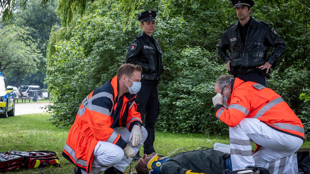 "Notruf Hafenkante - Dicke Fische": Dr. Haase (Fabian Harloff) und sein Kollege (Komparse) knien neben einem Mann (Jörg Malchow), der bewusstlos auf dem Rücken auf einer Wiese liegt. Die Polizistin Franzi Jung (Rhea Harder-Vennewald) und ihr Kollege Mattes Seeler (Matthias Schloo) stehen dahinter und beobachten nachdenklich die Umgebung.