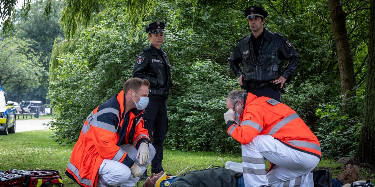 "Notruf Hafenkante - Dicke Fische": Dr. Haase (Fabian Harloff) und sein Kollege (Komparse) knien neben einem Mann (Jörg Malchow), der bewusstlos auf dem Rücken auf einer Wiese liegt. Die Polizistin Franzi Jung (Rhea Harder-Vennewald) und ihr Kollege Mattes Seeler (Matthias Schloo) stehen dahinter und beobachten nachdenklich die Umgebung.