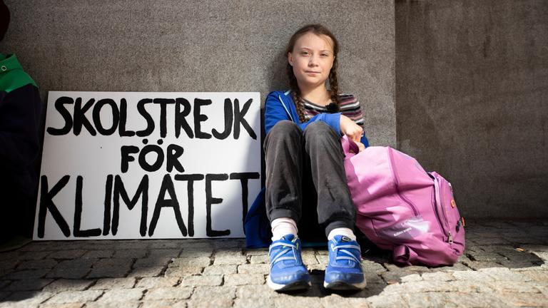 Die Klimaaktivistin Greta Thunberg sitzt neben einem Plakat mit der Aufschrift: "Skolstrejk för klimatet" (Schulstreik für das Klima).