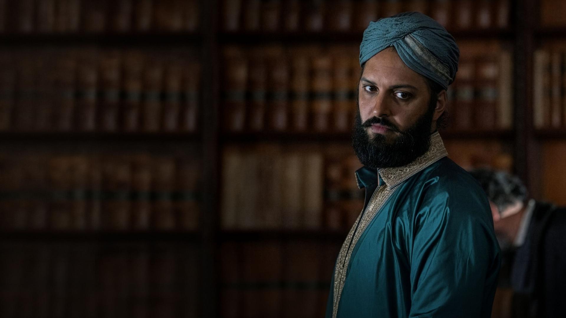 "Nautilus - Die Aktionäre": Nemo (Shazad Latif) steht in einem Raum vor einem Bücherregal. Er trägt elegante indische Kleidung und einen Dastar als Kopfbedeckung.
