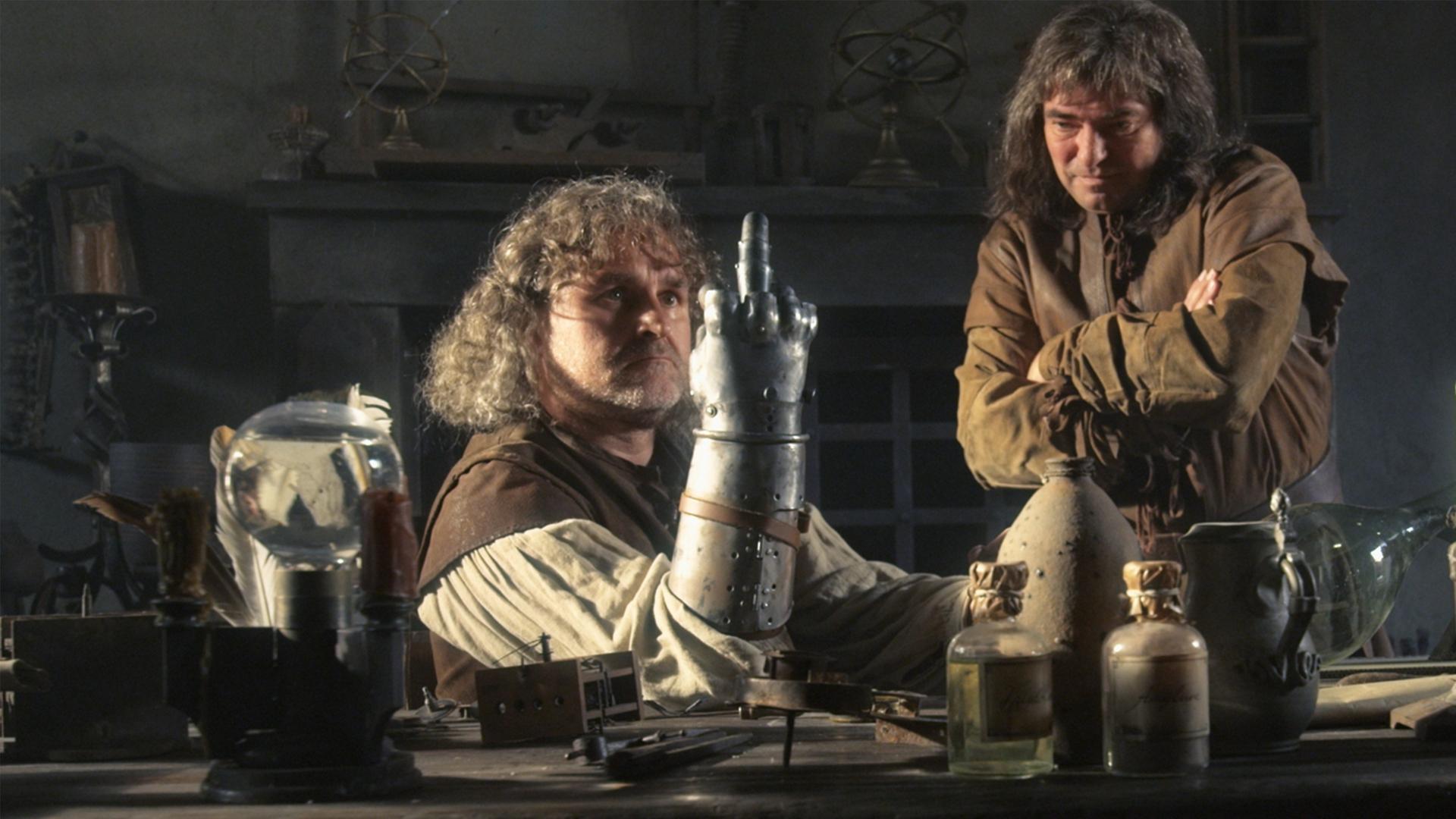 "Die glorreichen 10 - Die allerkrassesten Abstürze der Geschichte": Spielszene: Götz von Berlichingen sitzt an einer Werkbank, seinen rechten Arm mit der eisernen Hand auf die Werkbank gestützt. Von den Fingern dieser Hand ist nur der Mittelfinger ausgestreckt. Neben ihm steht ein Mann, der seine Arme vor der Brust verschränkt hat und nachdenklich auf die eiserne Hand schaut.