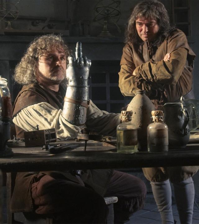 "Die glorreichen 10 - Die allerkrassesten Abstürze der Geschichte": Spielszene: Götz von Berlichingen sitzt an einer Werkbank, seinen rechten Arm mit der eisernen Hand auf die Werkbank gestützt. Von den Fingern dieser Hand ist nur der Mittelfinger ausgestreckt. Neben ihm steht ein Mann, der seine Arme vor der Brust verschränkt hat und nachdenklich auf die eiserne Hand schaut.