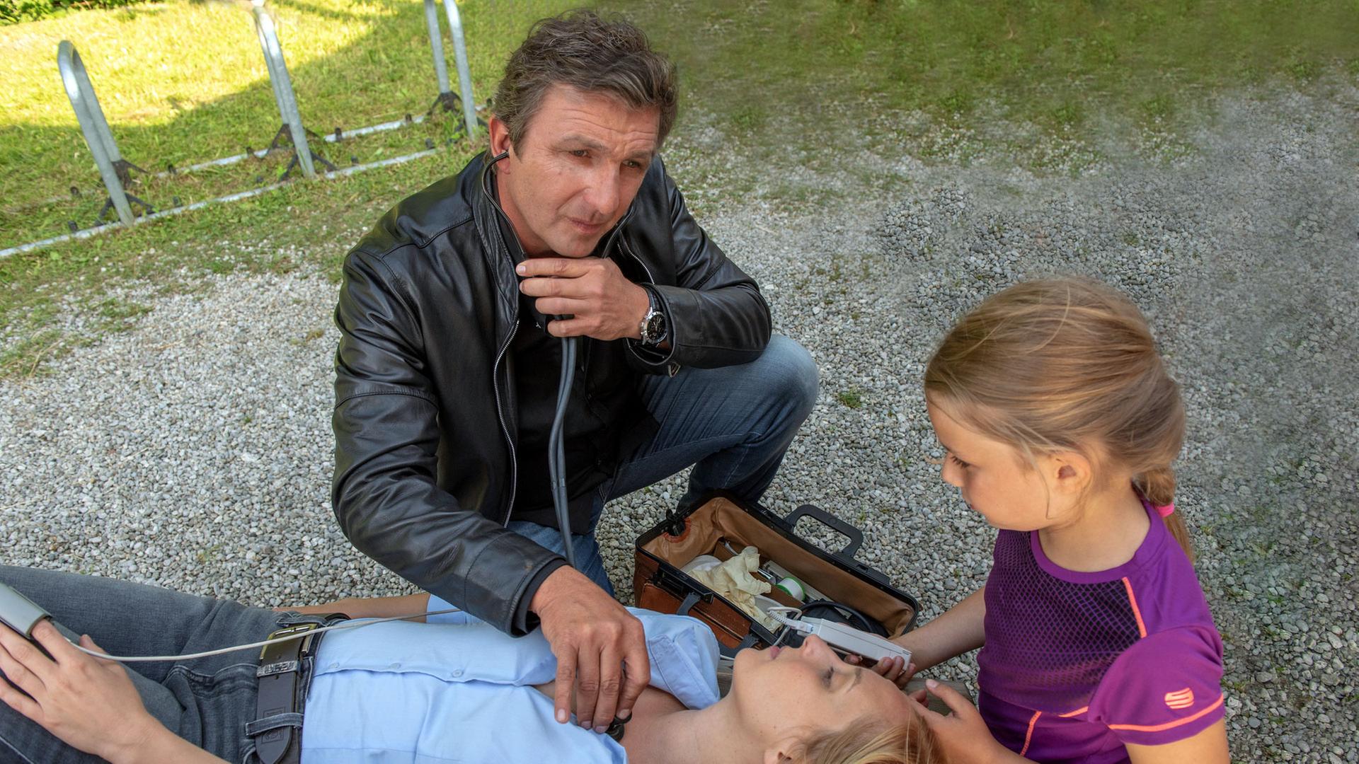 "Der Bergdoktor: Die Andere": Lena (Cordula Zielonka) ist zusammengebrochen und liegt auf einer Bank. Ihre Tochter Finja (Eve Marie Gleißner) sitzt daneben, Dr. Martin Gruber (Hans Sigl) hat sein Stethoskop im Einsatz.