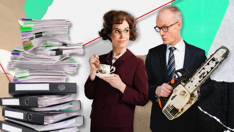 Maike Kühl und Max Uthoff stehen neben einem großen Stapel Aktenordner. Maike Kühl hält eine Tasse Tee in der Hand, Max Uthoff hält eine Motorsäge.