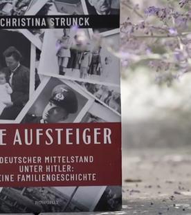 Draußen auf einer asphaltierten Straße steht vor lila Blütenzweigen ein Buch mit dem Titel "Die Aufsteiger" und alten Familienfotos auf dem Boden.