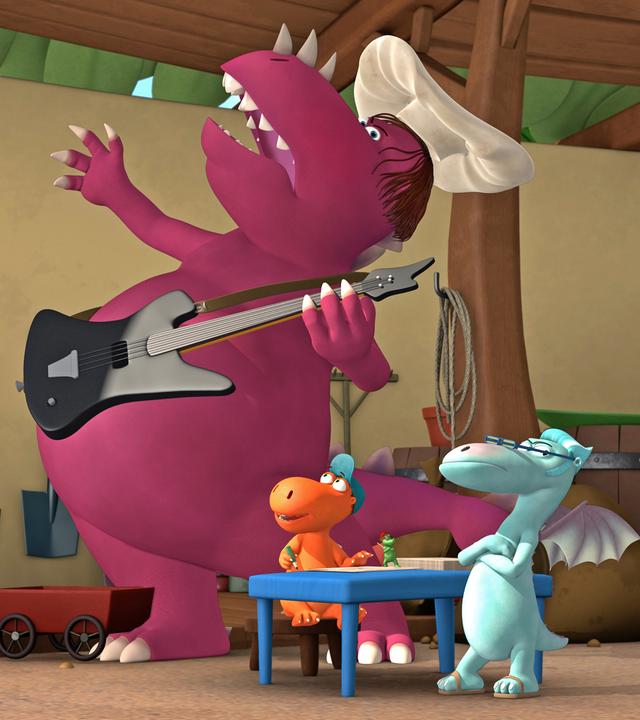 Der Chefkoch spielt E-Gitarre, während Kokosnuss begeistert zu ihm aufblickt. Ein blauer Drache steht mit verschränkten Armen daneben.