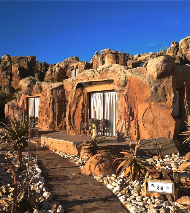 Inmitten von Felsen liegen drei Terrassen mit den Eingängen zu Höhlenzimmern. Davor ist ein Steingarten angelegt.