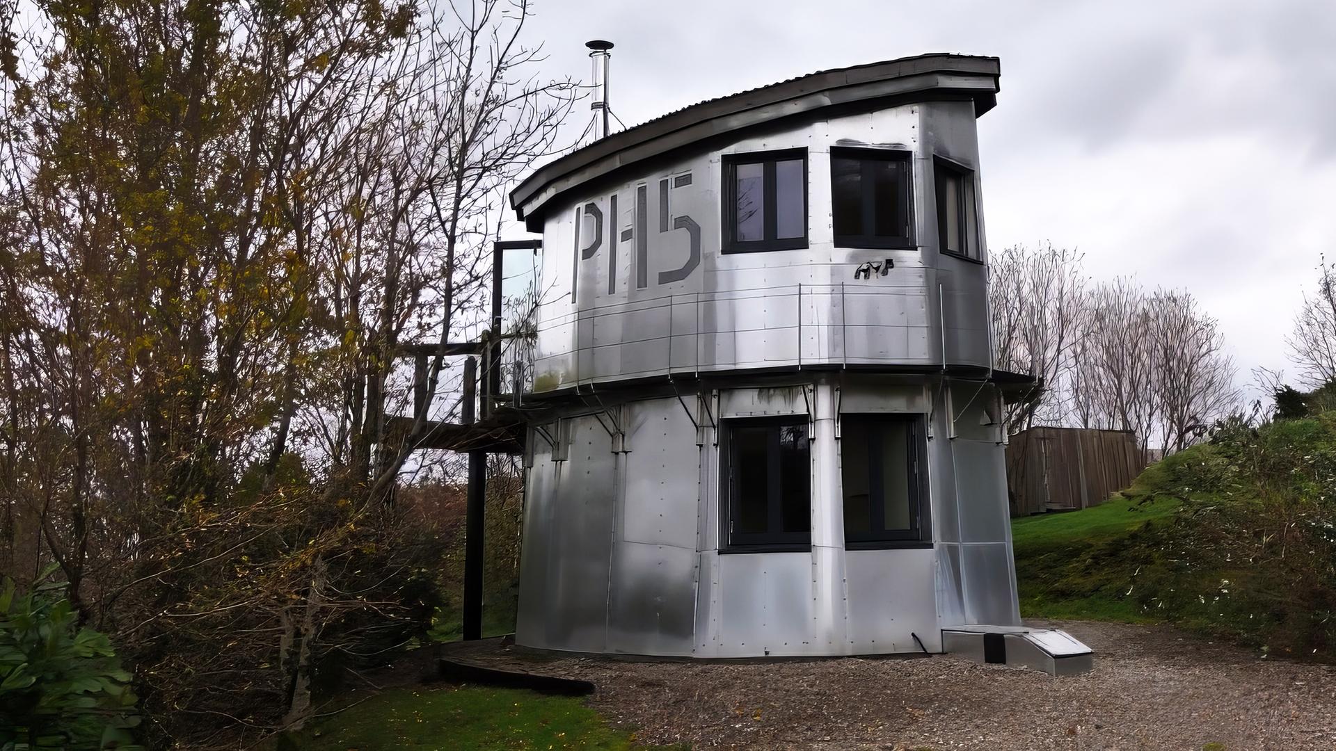 Ein silberner, zweistöckiger Wachturm mit großen Fenstern und der Nummer 'PH15' an der Seite steht an einem Weg im Grünen. Er ist umgeben von Bäumen und bewölktem Himmel