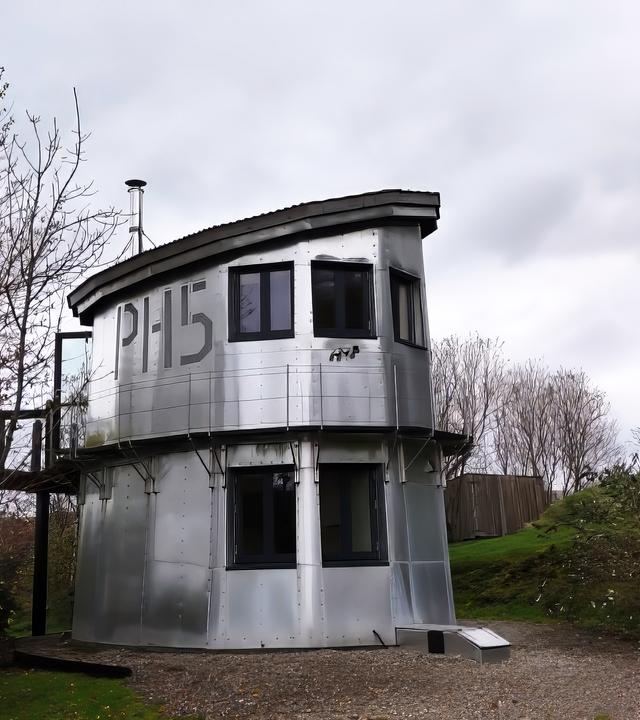 Ein silberner, zweistöckiger Wachturm mit großen Fenstern und der Nummer 'PH15' an der Seite steht an einem Weg im Grünen. Er ist umgeben von Bäumen und bewölktem Himmel