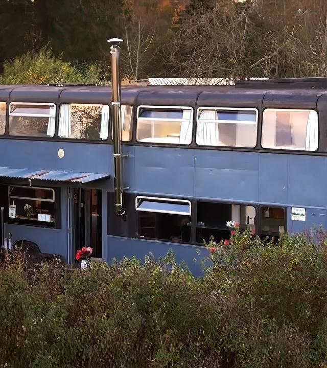 Ein blauer Doppeldeckerbus wurde in ein Haus umgebaut. Er steht im Grünen und hat mehrere Fenster, einem Schornstein und einen überdachten Eingang.