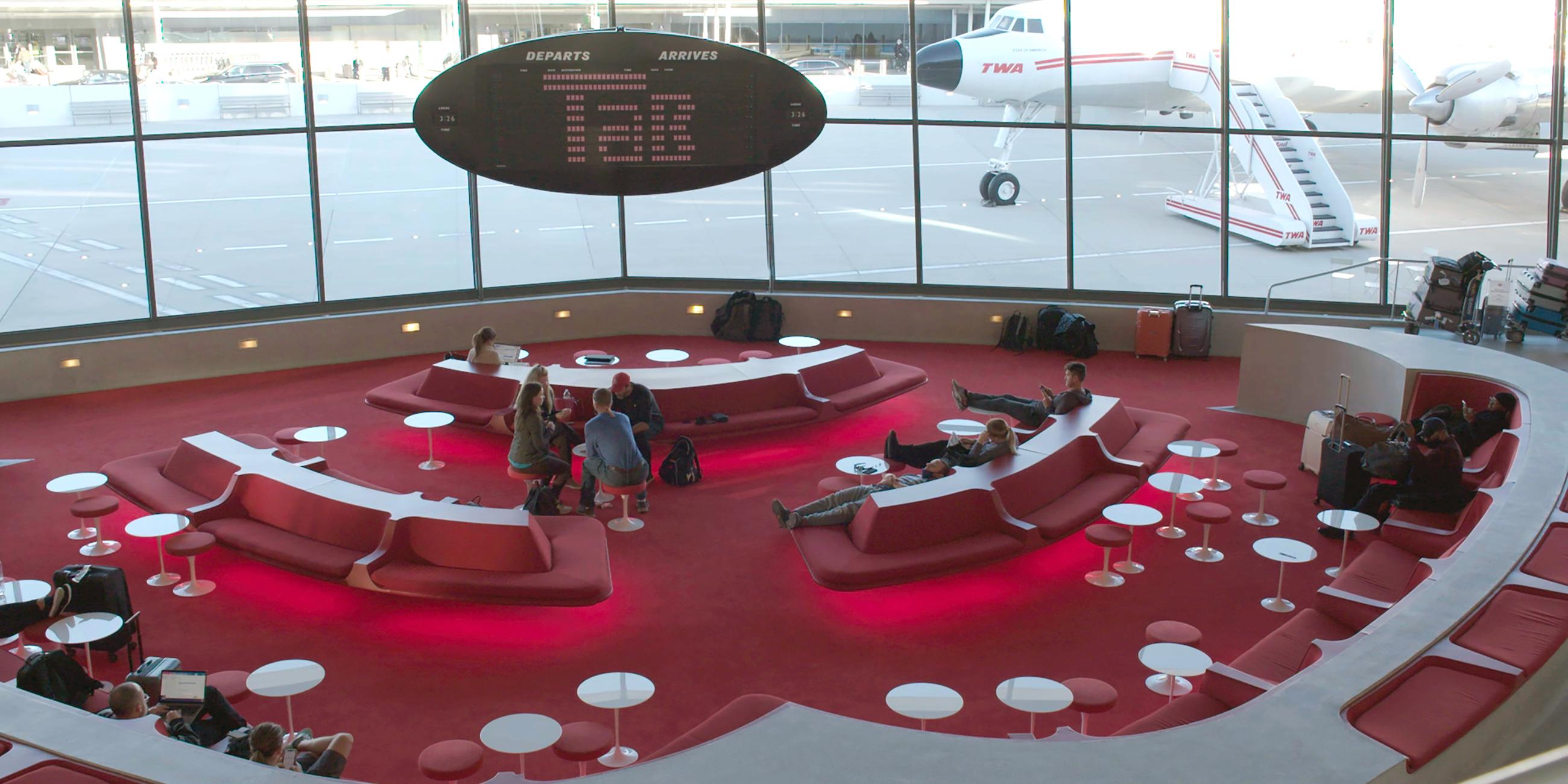 Mehrere Personen sitzen in der Wartehalle eines Flughafenterminals. Hinter der Fensterfront steht ein Flugzeug.
