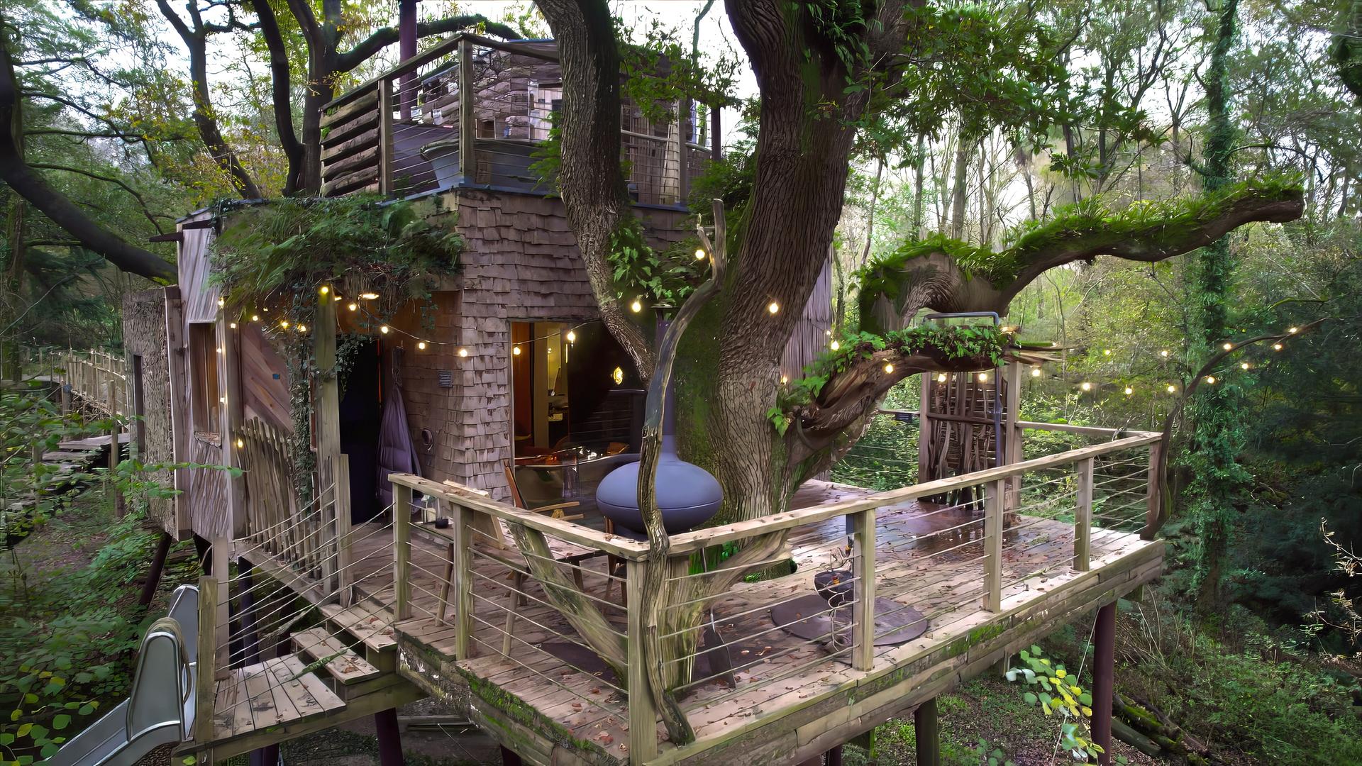 In einem großen Baum zwischen mehreren Ästen befindet sich ein Baumhaus mit Holzgeländer und Lichterkette, umgeben von weiteren Bäumen.