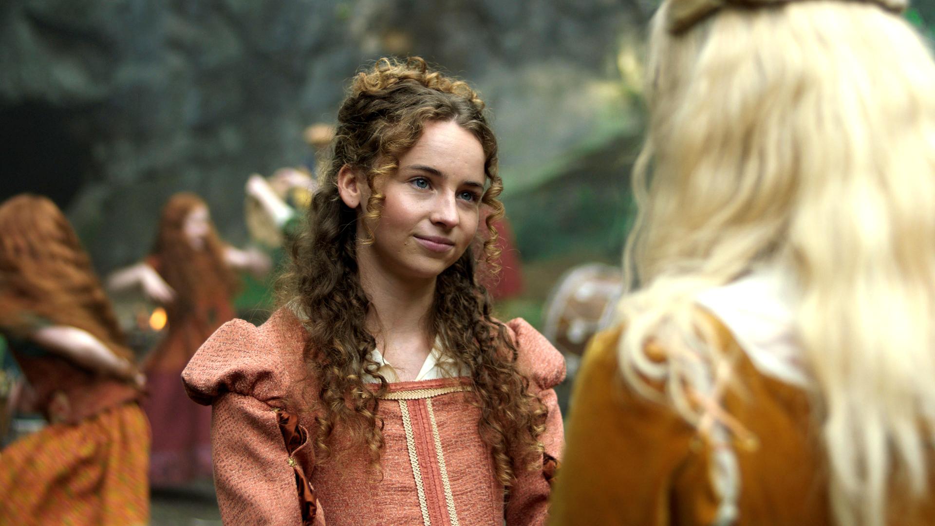 Im 16. Jahrhundert draußen stehen sich die Schwestern Maggie und Elinor in ihren edlen Kleidern gegenüber. Maggie schaut ernst aber liebevoll mit leicht geneigtem Kopf zu Elinor, die nur von hinten zu sehen ist. Der Hintergrund ist unscharf, doch es sind tanzende Kinder in rostroten Kleidern zu erkennen.