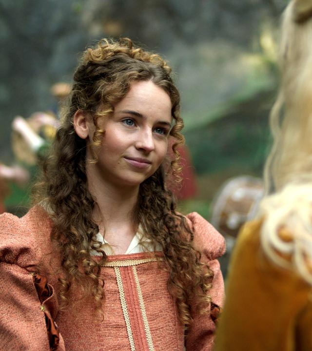 Im 16. Jahrhundert draußen stehen sich die Schwestern Maggie und Elinor in ihren edlen Kleidern gegenüber. Maggie schaut ernst aber liebevoll mit leicht geneigtem Kopf zu Elinor, die nur von hinten zu sehen ist. Der Hintergrund ist unscharf, doch es sind tanzende Kinder in rostroten Kleidern zu erkennen.