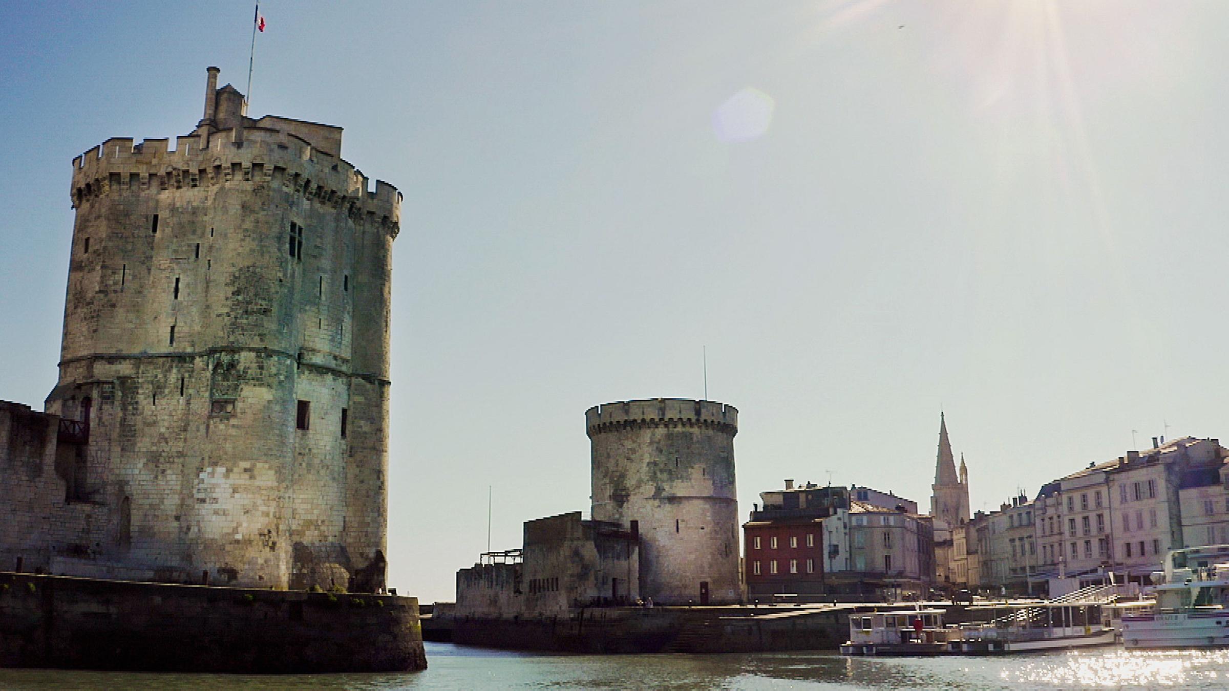 "Die Belagerung von La Rochelle": An der Hafeneinfahrt einer Stadt stehen zwei Türme. Im Hintergrund sind bunte Häuser zu sehen.