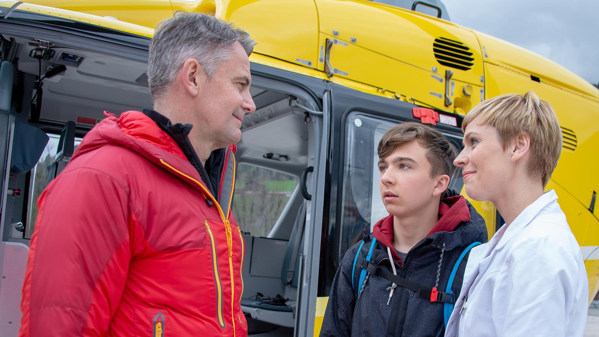 Dr. Verena Auerbach (Gundula Niemeyer) steht mit Leon (Tim Marco Rietz) am Rettungshubschauer Michael (Robert Lohr) gegenüber. Verena und Michael lächeln Leon an.