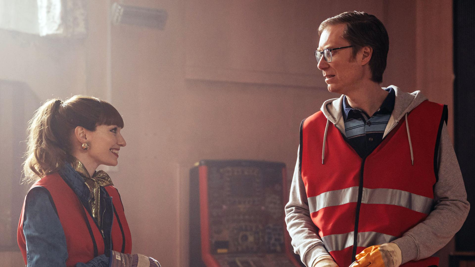 "The Outlaws - Die Bootsparty": Lady Gabby (Eleanor Tomlinson) und Greg (Stephen Merchant) stehen in einem vermüllten, dunklen Gebäude nebeneinander und schauen sich fröhlich an. Dabei tragen beide orangene Warnwesten.