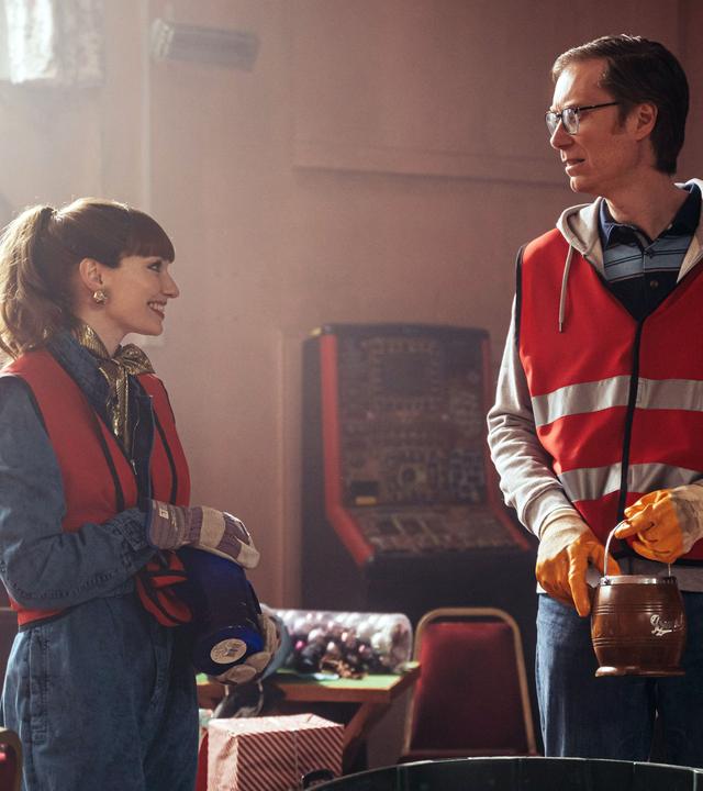 "The Outlaws - Die Bootsparty": Lady Gabby (Eleanor Tomlinson) und Greg (Stephen Merchant) stehen in einem vermüllten, dunklen Gebäude nebeneinander und schauen sich fröhlich an. Dabei tragen beide orangene Warnwesten.