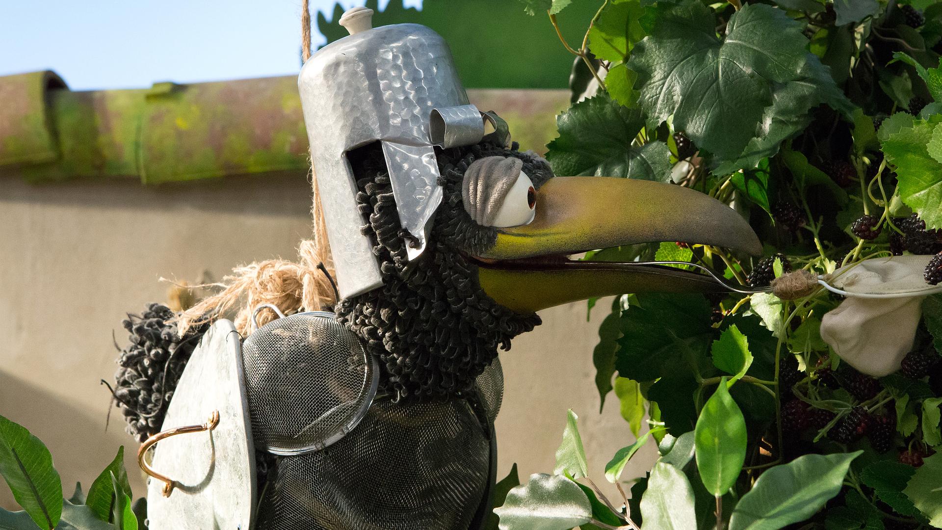 Rudi trägt ein Ritterkostüm und hängt an einem Seil. Er versucht, mit einem Kescher Brombeeren vom Strauch zu pflücken. 