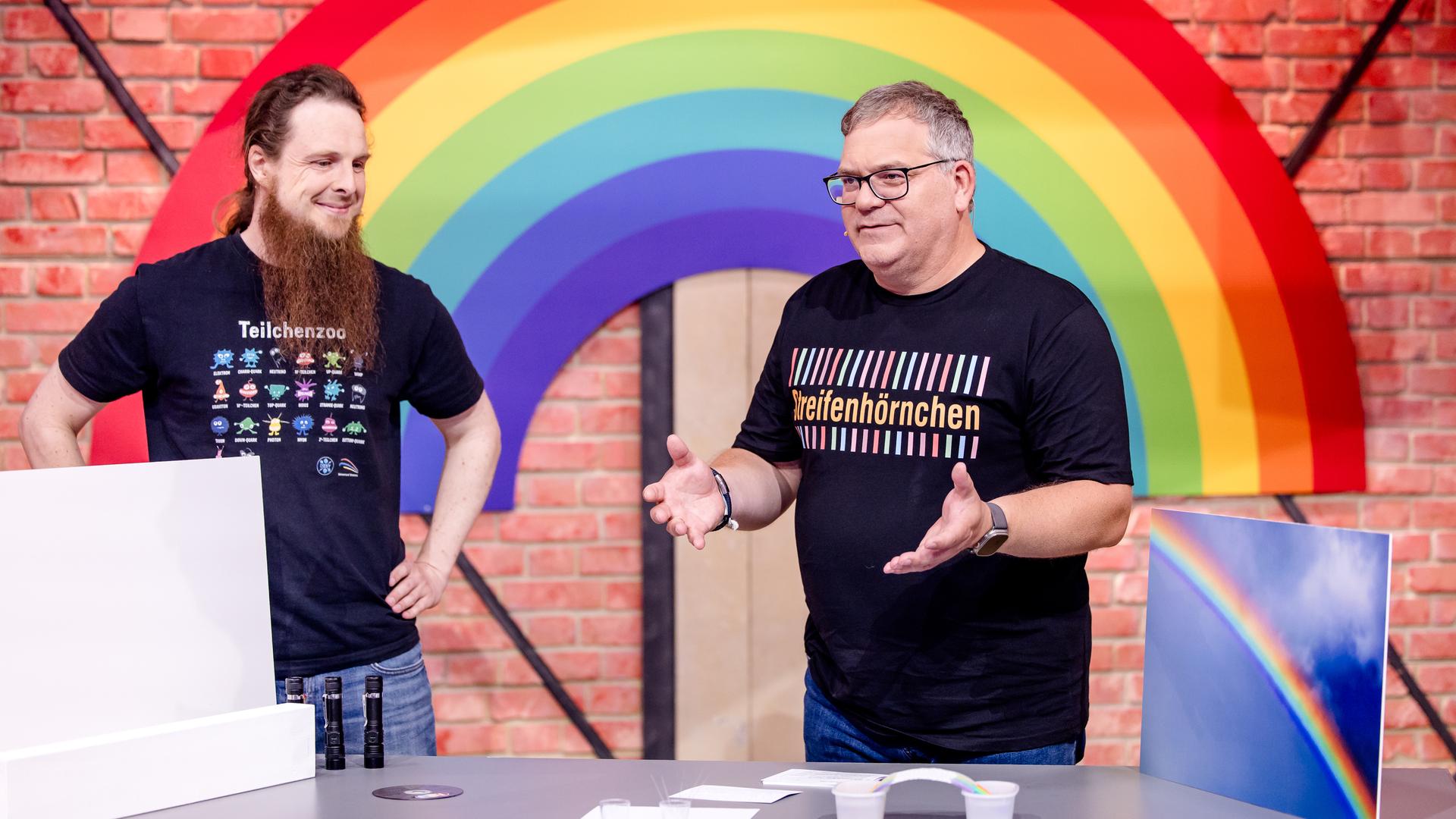 "1, 2 oder 3 - Die bunte Welt der Streifen": Michael Büker und Elton stehen im Studio hinter einem Tisch. Hinter ihnen ist ein großer Regenbogen zu sehen.