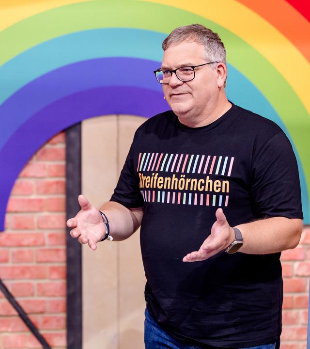 "1, 2 oder 3 - Die bunte Welt der Streifen": Michael Büker und Elton stehen im Studio hinter einem Tisch. Hinter ihnen ist ein großer Regenbogen zu sehen.