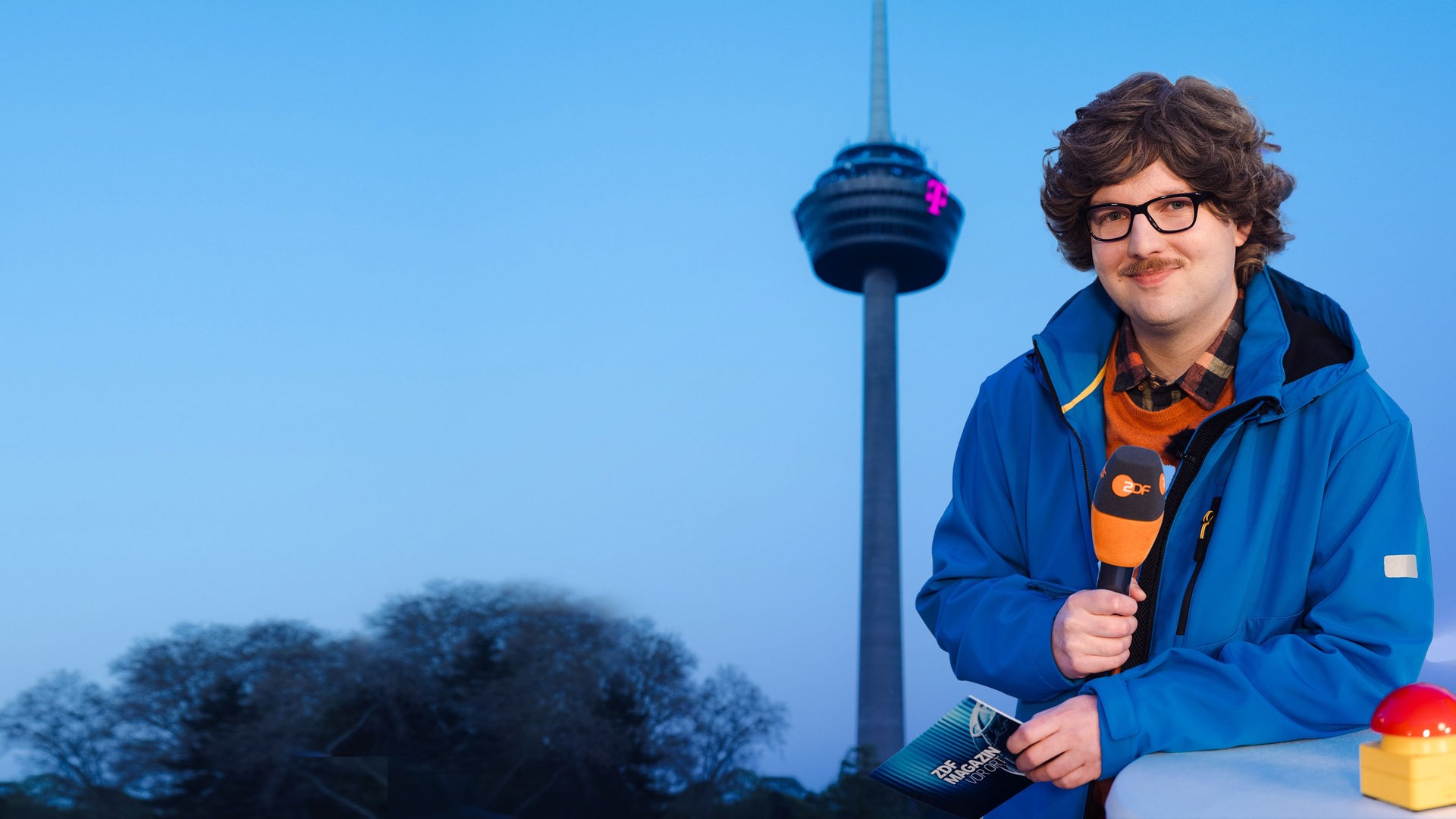 Reporter Peter-Stefan Thomas (Chris Sommer) steht vor dem Kölner Fernsehturm Colonius und hält ein ZDF Mikrofon in der Hand.