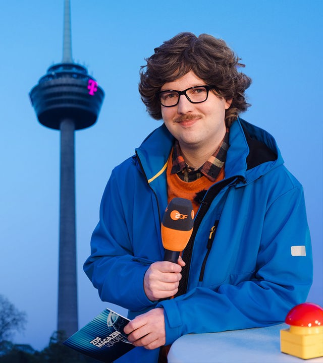 Reporter Peter-Stefan Thomas (Chris Sommer) steht vor dem Kölner Fernsehturm Colonius und hält ein ZDF Mikrofon in der Hand.