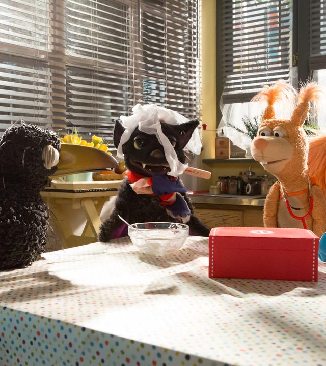 Rabe Rudi, Fledermaus Loulou, die in Mullbinden gehüllt ist und ein Fieberthermometer im Maul hat, und Eichhörnchen Ella hocken auf dem Küchentisch. Rudi und Ella schauen besorgt auf die vermeintliche Vampi.