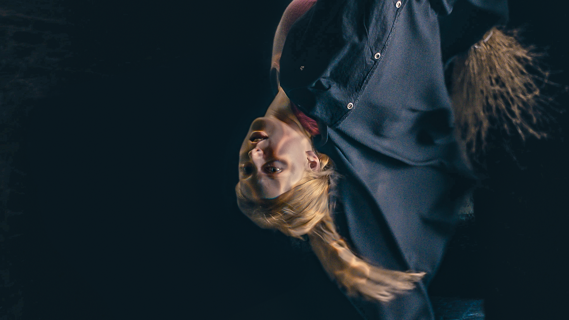 Mildred in einem schwarzen Cape fliegt auf einem Besen kopfüber vor einem schwarzen Hintergrund - es ist ein Tunnel. Sie hat einen Schimmer auf ihrem Gesicht und ihrem langen rot-blonden Haar - das Zeichen, das gerade ein Zauber geschieht.