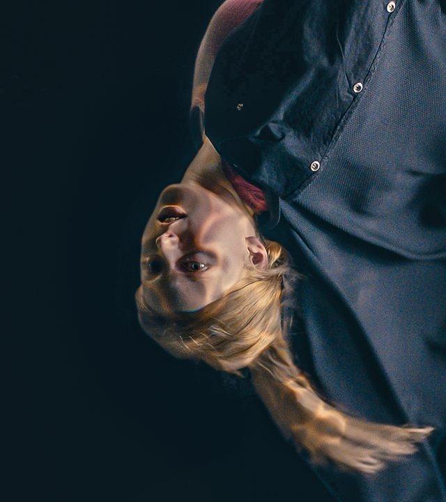 Mildred in einem schwarzen Cape fliegt auf einem Besen kopfüber vor einem schwarzen Hintergrund - es ist ein Tunnel. Sie hat einen Schimmer auf ihrem Gesicht und ihrem langen rot-blonden Haar - das Zeichen, das gerade ein Zauber geschieht.