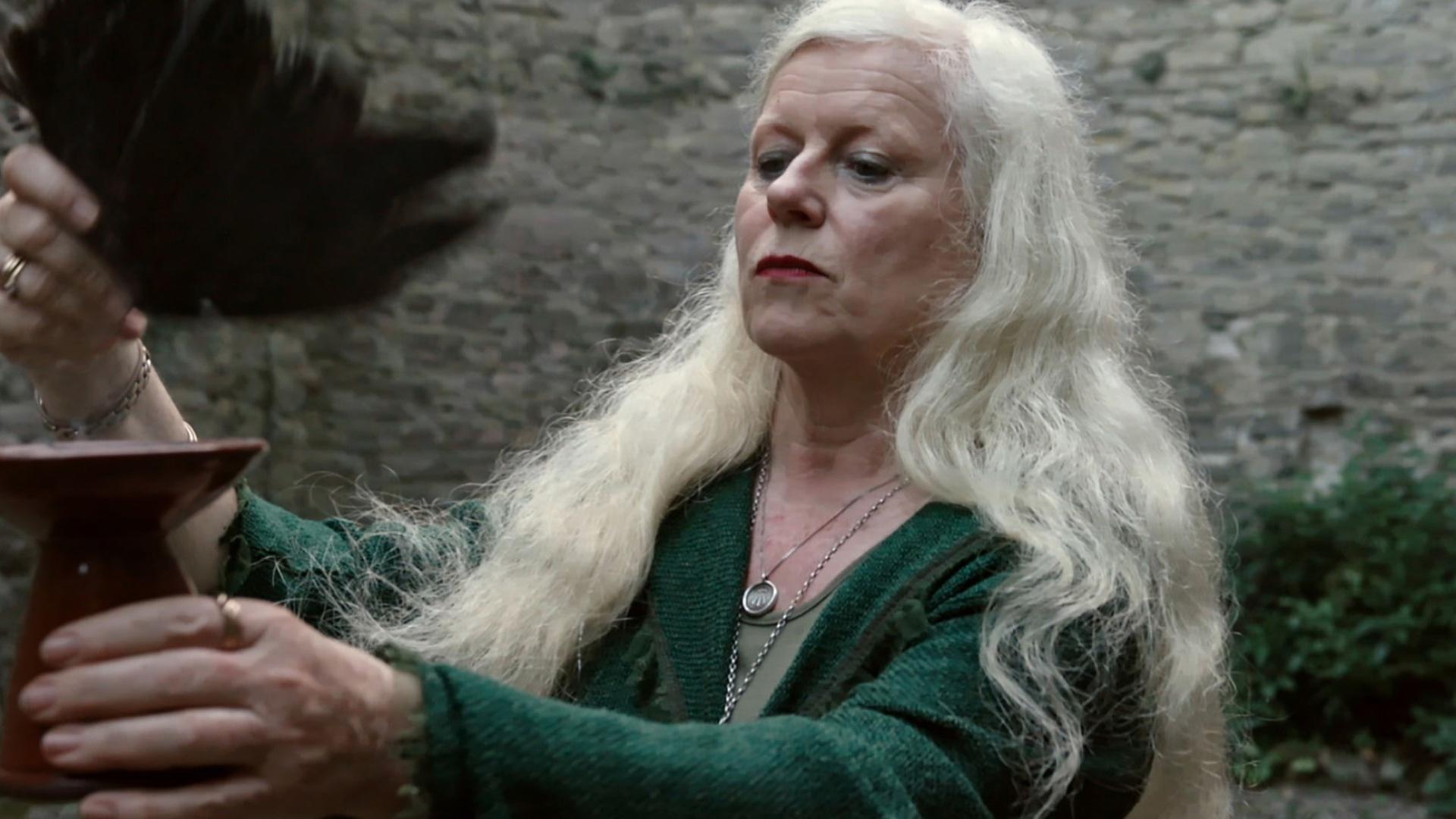 Eimear Burke mit langen, grau-blonden Haaren hält einen Kelch in der einen Hand und eine schwarze Feder in der anderen.